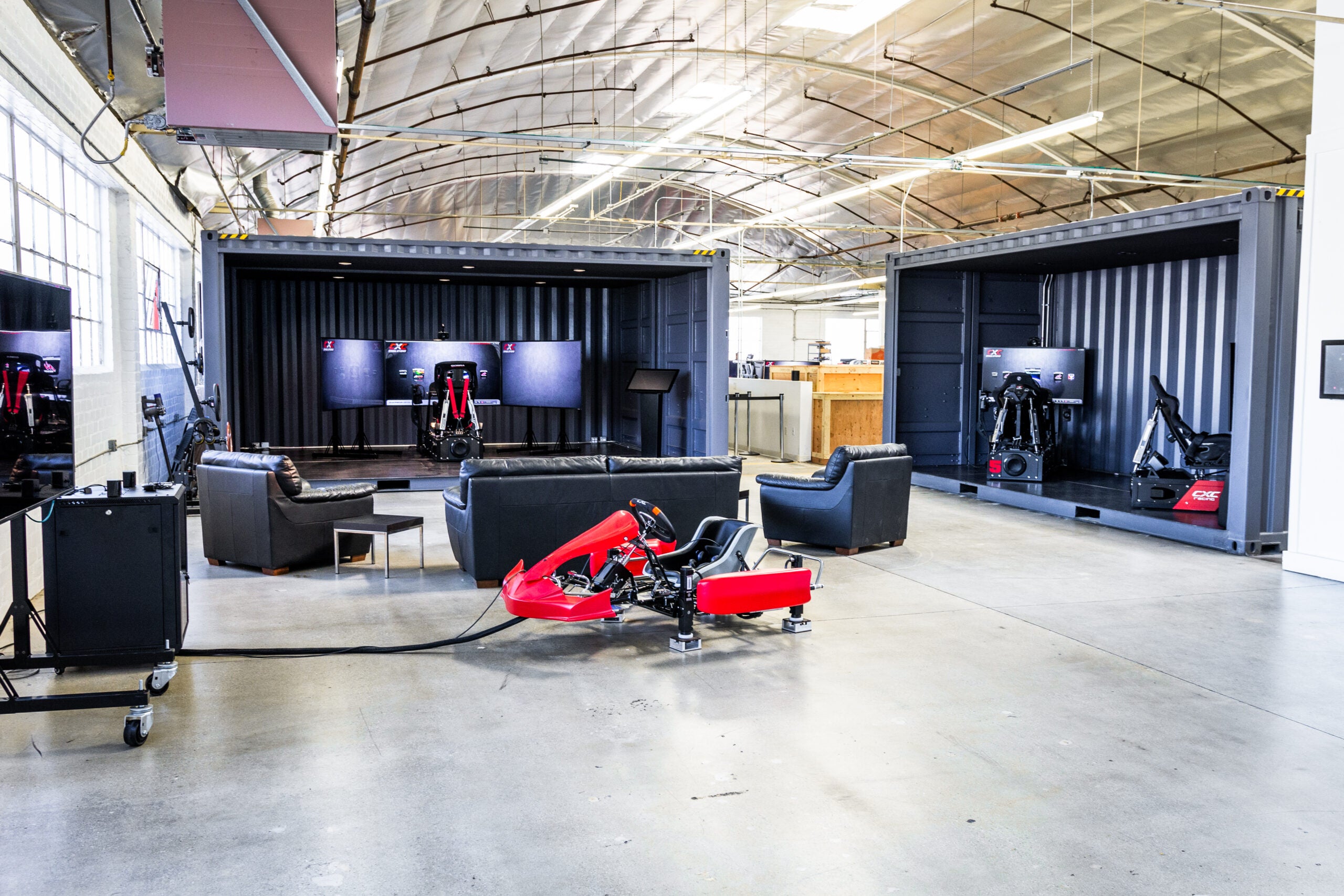A brightly lit warehouse interior with two half opened intermodal containers within it. Inside of the containers are driving simulators. In the foreground, some couches and a kart make up the decor.