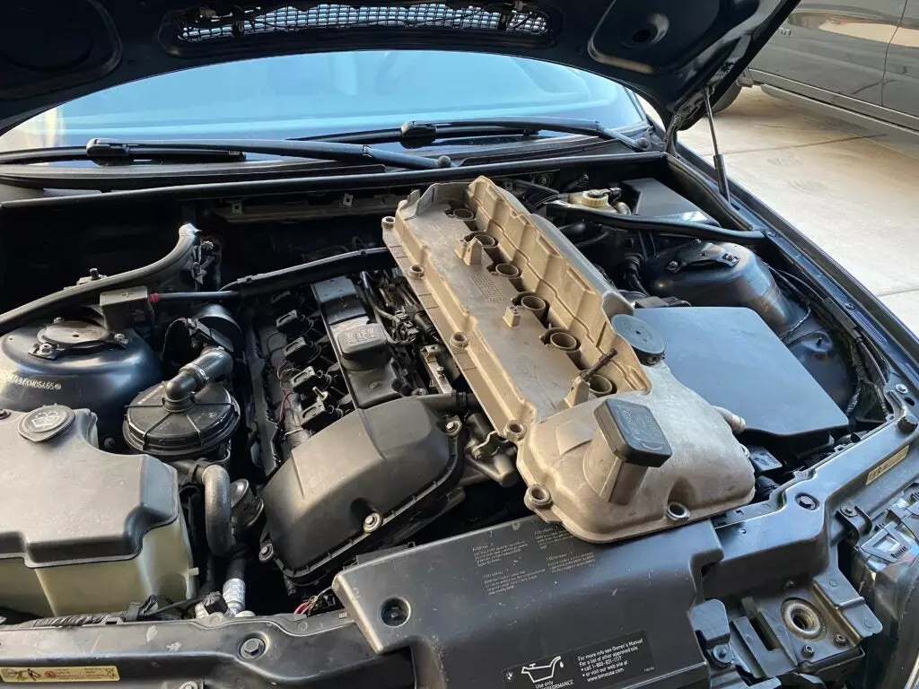 An aluminum M56 valve cover next to a normal plastic M54 valve cover in the engine bay of a 2004 BMW 330i.