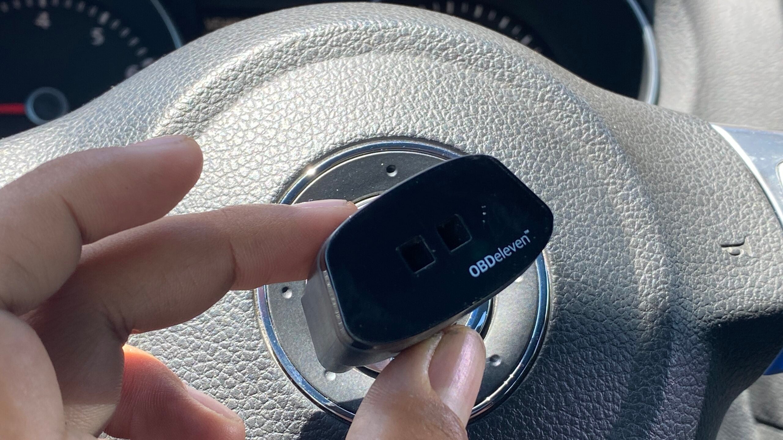 An OBDEleven bluetooth dongle held by a hand. A 2010 VW GTI steering wheel is in the background.