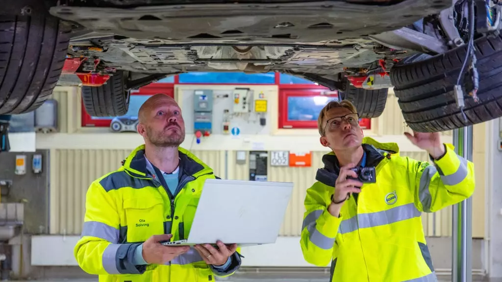 Two Volvo employees inspect the underside of a wrecked vehicle on a lift.