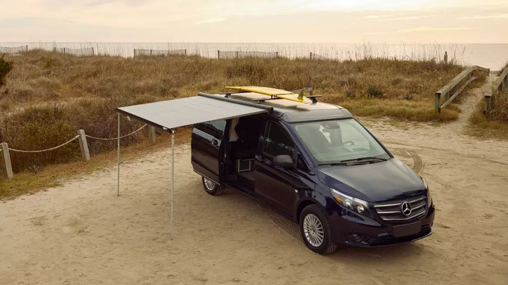 A Mercedes-Benz camper van with a yellow surfboard on top and a canopy.