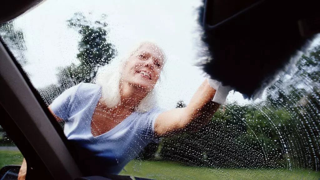An older woman washes a car windshield with a microfiber mitt.